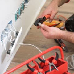 An individual is partially rewiring electrical cables, expertly plugging a connector into the panel. The focus remains sharp, dressed in a red and black outfit. A red toolbox with pliers and other tools rests on the wooden floor, ensuring everything needed for landlord safety certificates is at hand.